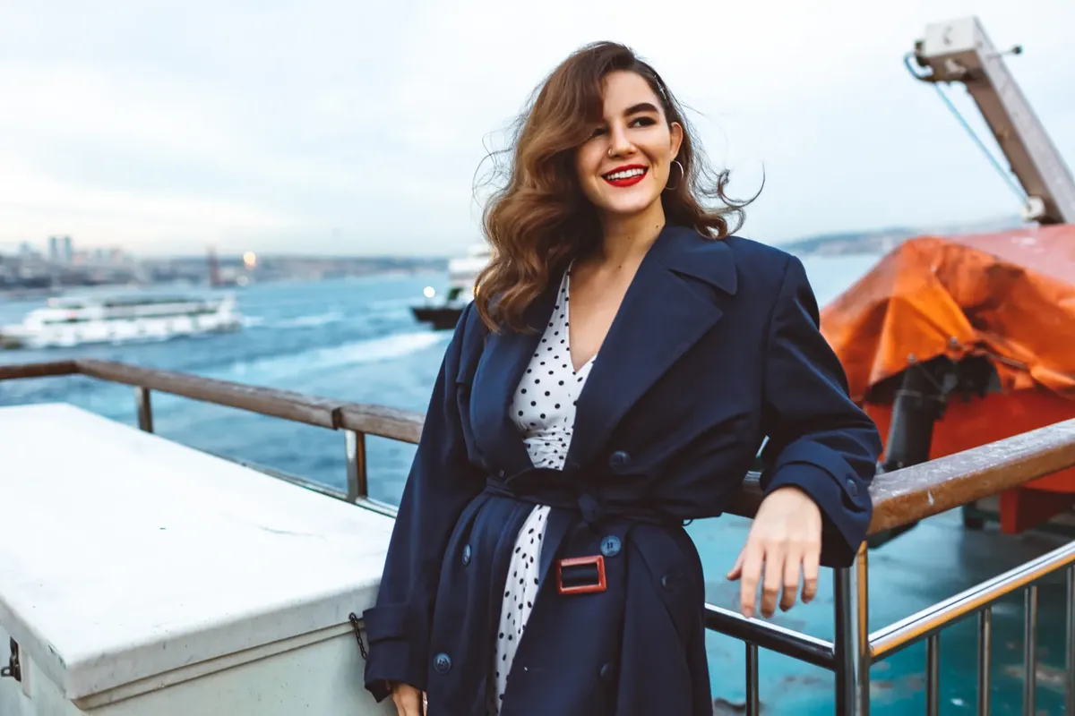 Fashionable woman in a blue coat smiles by the sea, evoking a sense of joy and style.