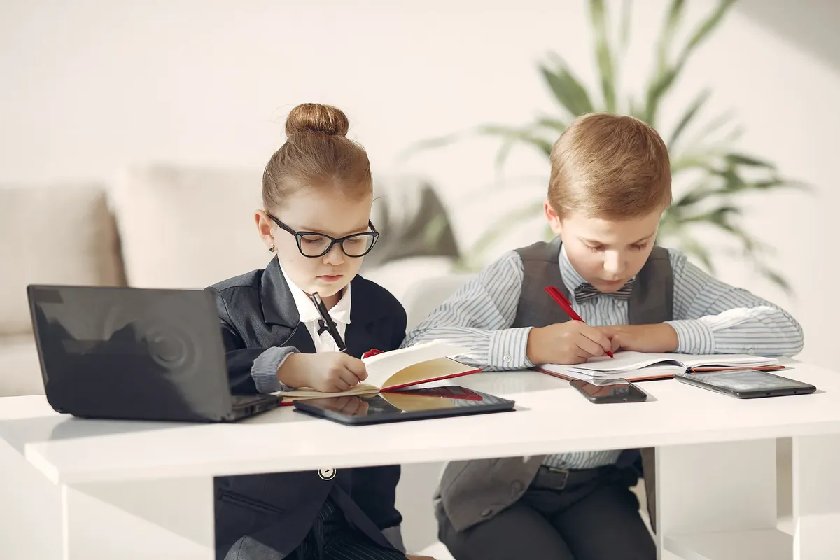 Cute business children in uniform using laptop and writing information in planners while making up startup strategy in modern workspace