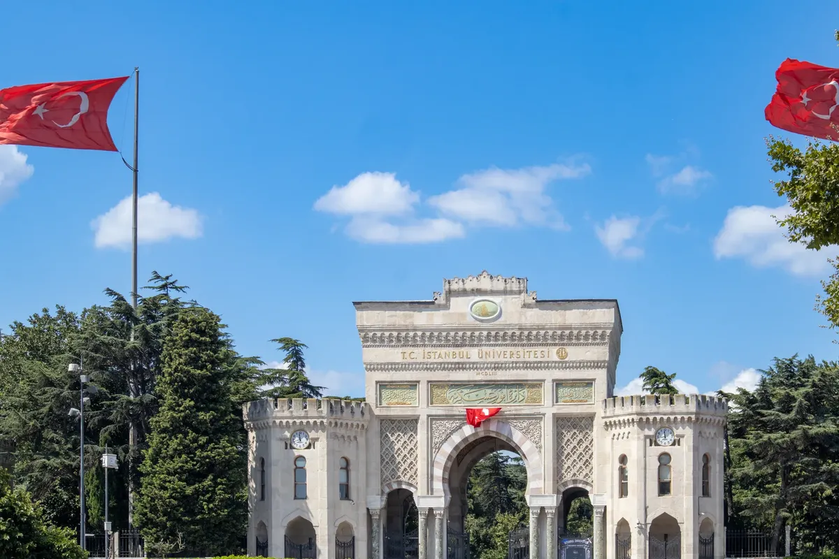 The iconic entrance gate of Istanbul University featuring Turkish flags and lush greenery.