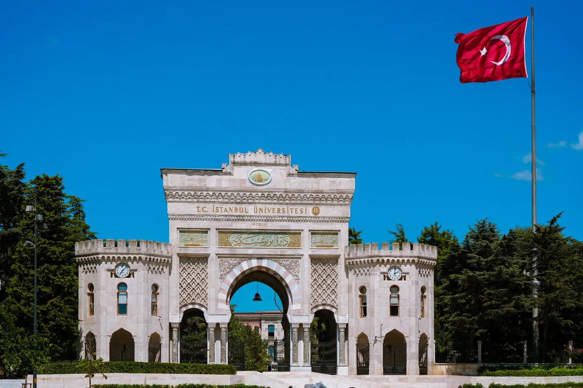 The grand entrance of Istanbul University adorned with a Turkish flag under a clear blue sky.