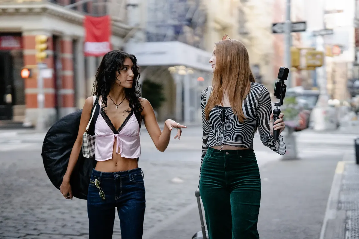 Two young women walking through city streets with luggage, vlogging their travel experience.